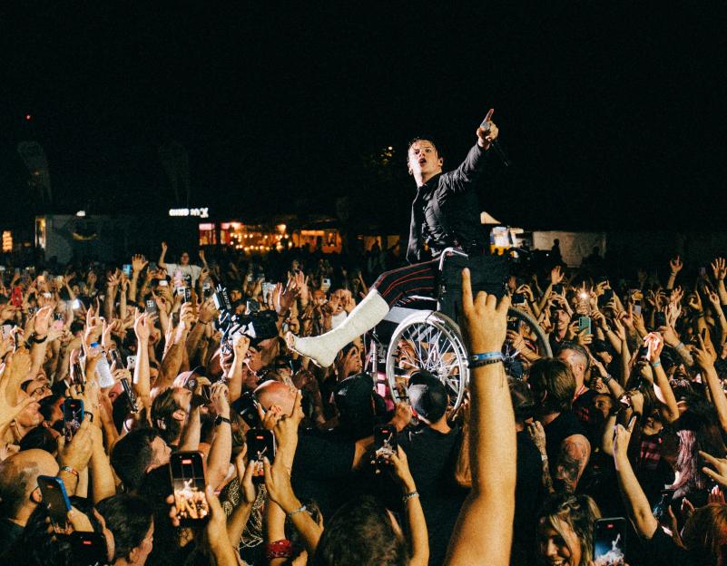 Yungblud on stage in a wheelchair with a cast on his leg, pointing at the crowd while they raise their hands and phones.