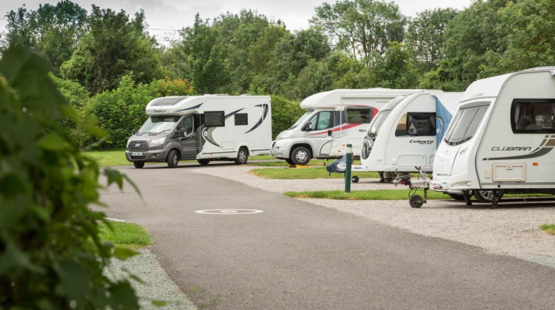 Campsite with several RVs and travel trailers parked on gravel beside an asphalt road, surrounded by trees.