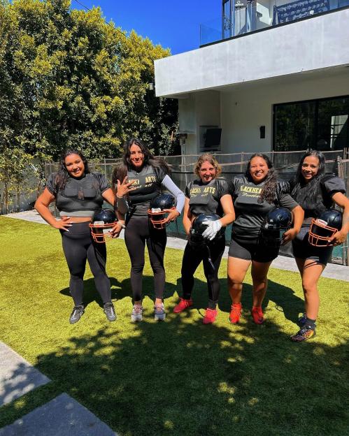 Five women in football gear holding helmets in a yard.