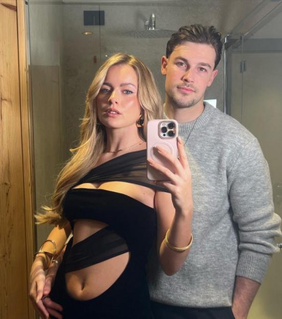 A woman in a black cutout dress taking a mirror selfie with a man in a gray sweater.
