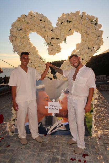Two men standing in front of a heart-shaped floral arch and a "Will you marry me?" puzzle photo.