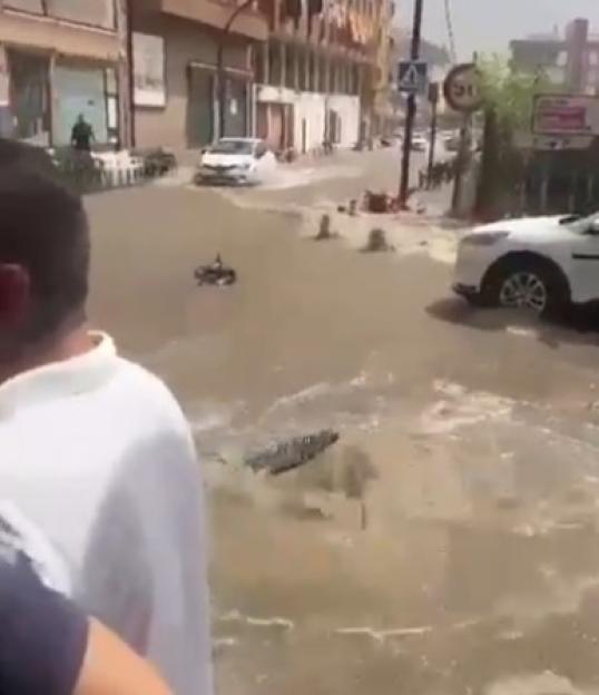 A flooded street in a city in Spain, with cars partially submerged and debris floating in the muddy water.