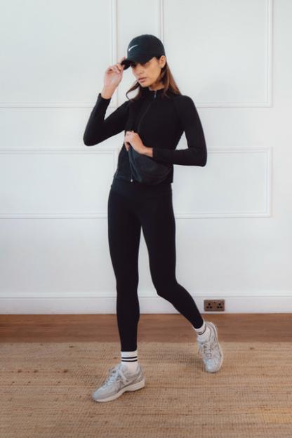 A woman in a black Nike cap, black long-sleeved athletic top, black leggings, and white sneakers with white socks.
