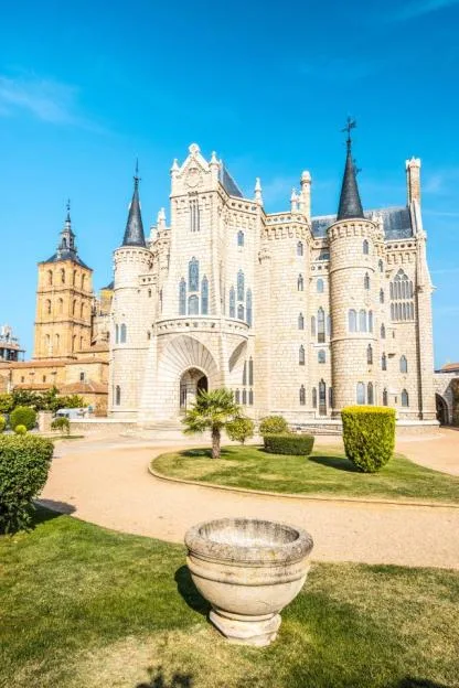 The Episcopal Palace of Astorga by Antoni Gaudí, a stone palace with multiple turrets, stained-glass windows, and Gothic architectural details, with a large, older church tower behind it to the left.