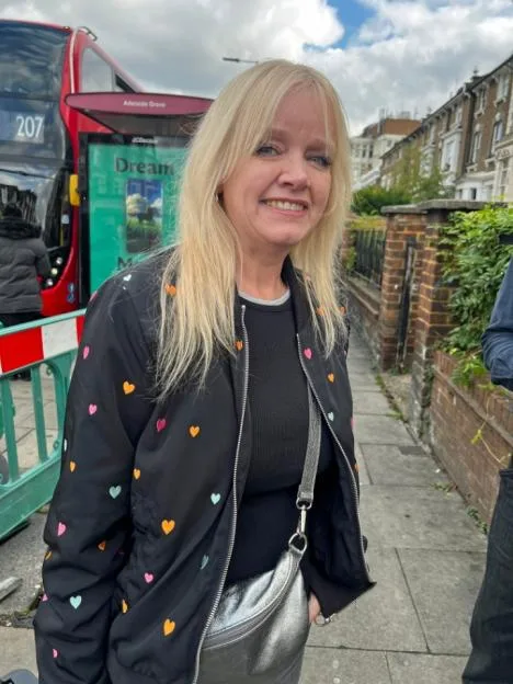 A blonde woman smiling on Uxbridge Road in London, with a red double-decker bus in the background.