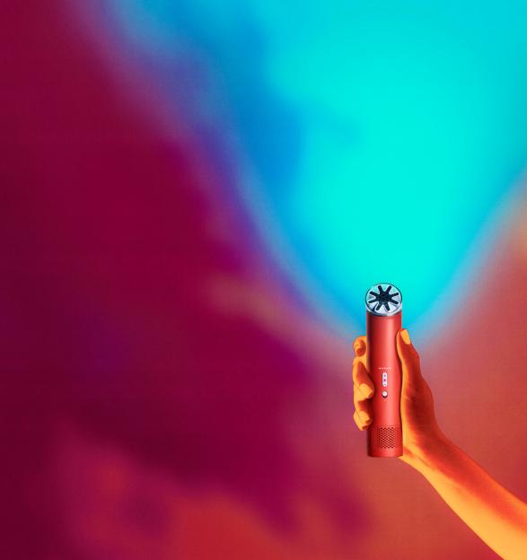 Hand holding a red device with a silver head resembling a fan, against a background of red, orange, and blue hues.