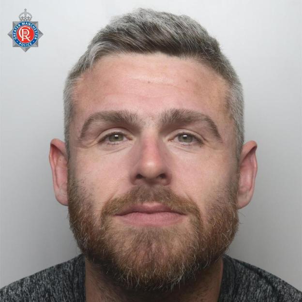 Mugshot of John Colin Spiby, a man with graying hair and a beard, wearing a gray shirt, with the Greater Manchester Police emblem in the top left corner.