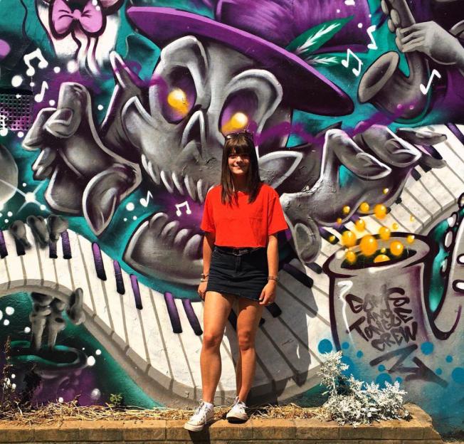 A young woman in a red shirt and black skirt stands in front of a colorful graffiti mural featuring a skull wearing a purple hat.