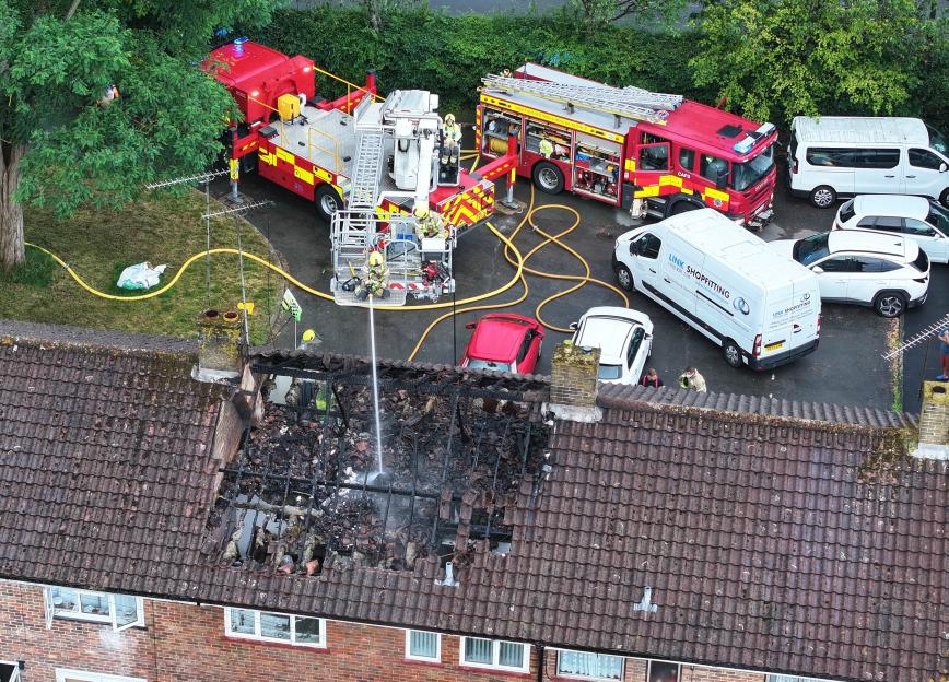 Smoke and flames rise from the roof of a house at night, next to another house with a tall, pointed roof.