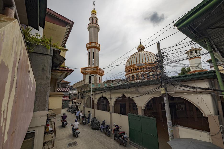 A mosque with a tall minaret and a large dome seen from a narrow street lined with buildings and parked motorcycles in Davao City, Philippines.