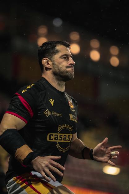 A Catalans Dragons player in a black and red uniform with yellow accents, looking to his right on a dark field.