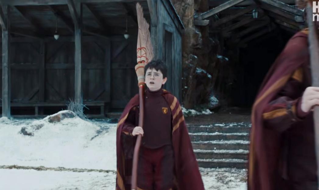 Harry Potter wearing Quidditch robes and holding a broomstick, standing in the snow in front of wooden buildings.
