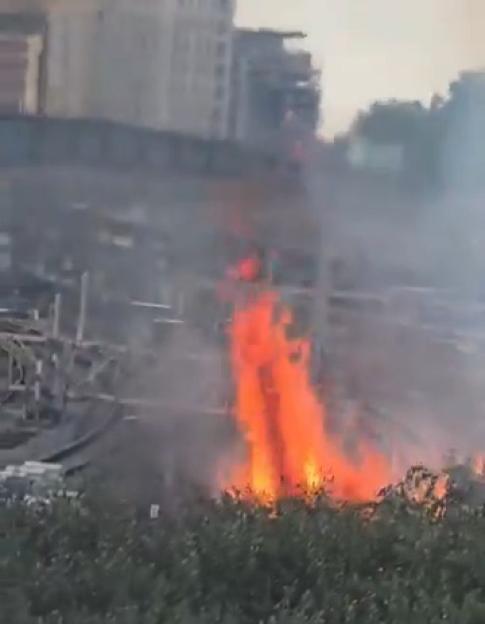 A fire burns near a railway line and shrubbery with buildings in the background.