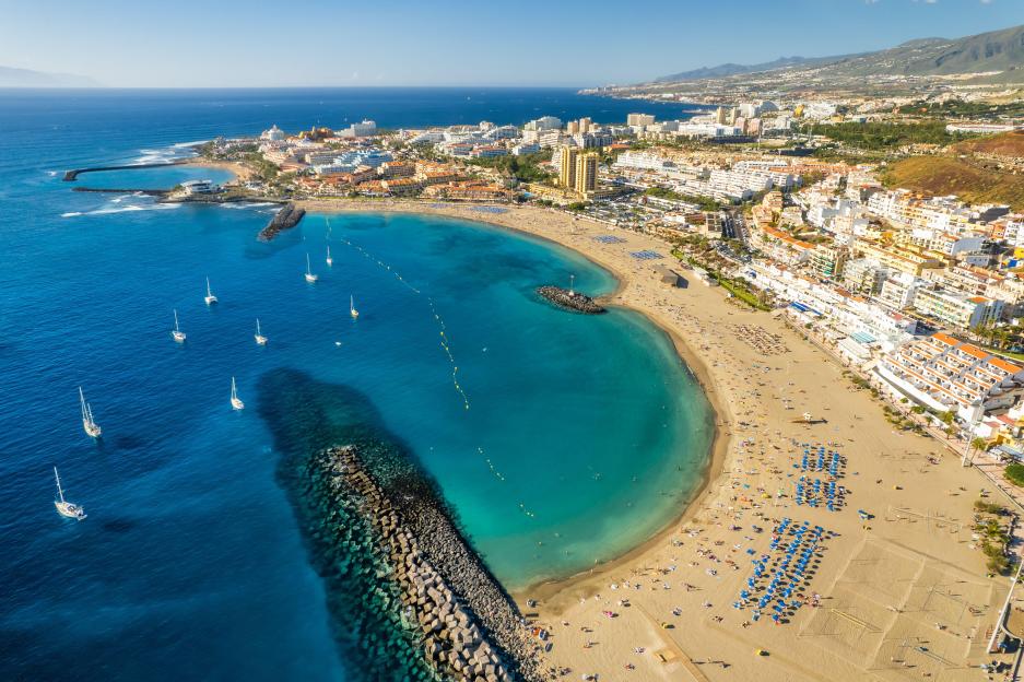 Aerial view of Playa de Las Vistas beach in Los Cristianos, Tenerife, with yachts in the ocean and hotels along the coastline.