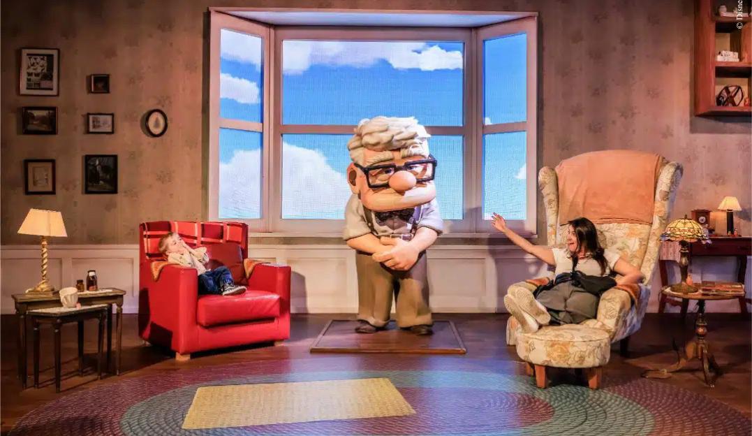 Mundo Pixar immersive experience with a large model of Carl Fredricksen from Up, two people in armchairs, and a window displaying a sky with clouds.