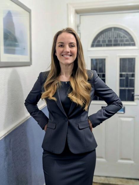 A smiling woman in a dark suit with her hands on her hips, standing in a hallway.