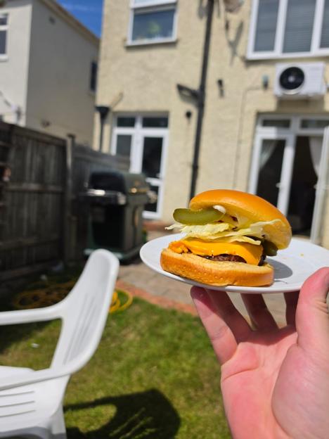 Hand holding a cheeseburger with pickles and lettuce on a plate in a backyard with a grill.