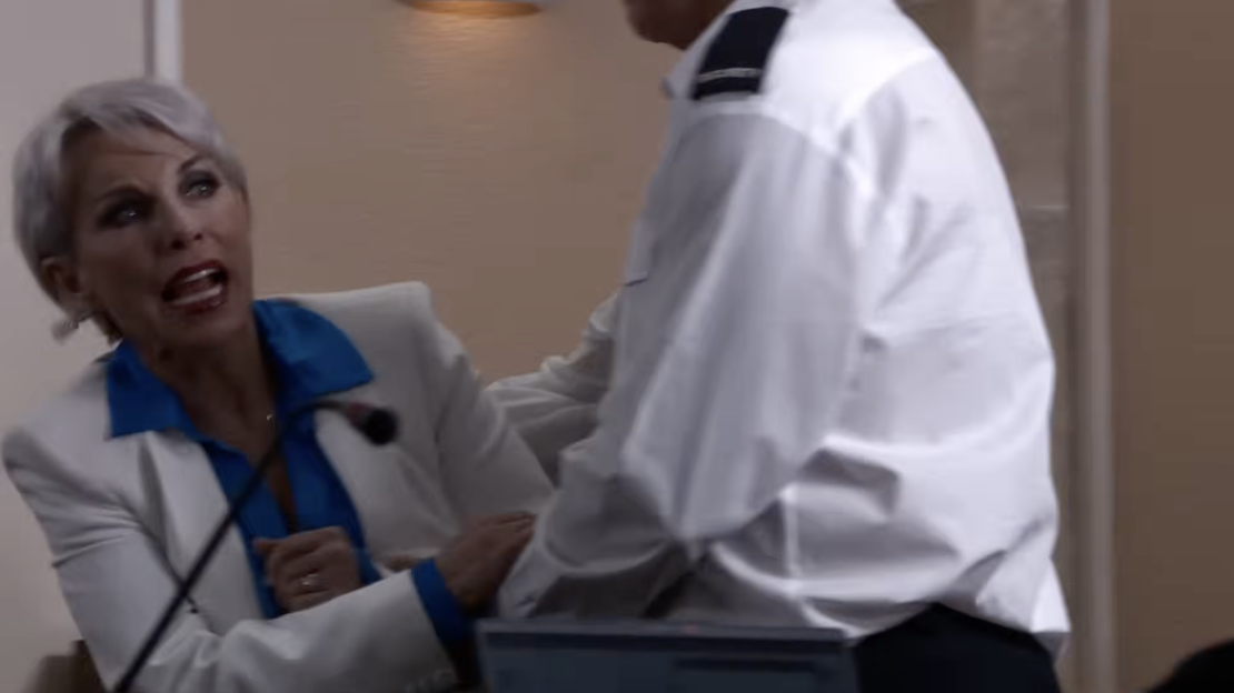 A woman with gray hair, wearing a white blazer and blue shirt, shouting with a microphone in front of her, with a man in a white uniform standing next to her.