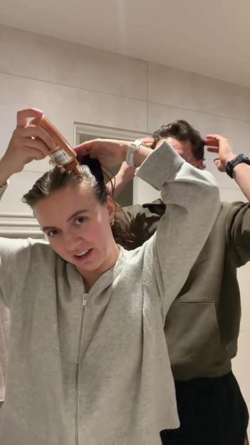 A woman applying hair product to her scalp, with a man in the background.