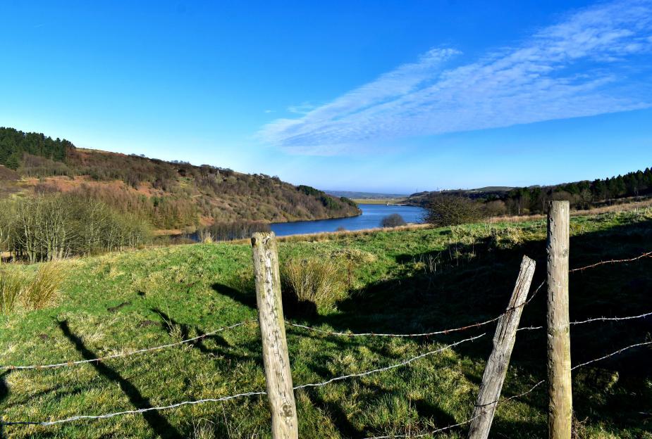 Scammonden Water reservoir and M62 motorway, seen through a barbed wire fence from a grassy hillside.