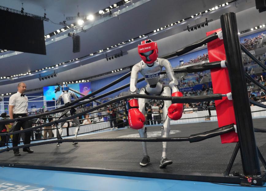 A humanoid robot in red boxing gear stands in a boxing ring, with another robot and a referee in the background.