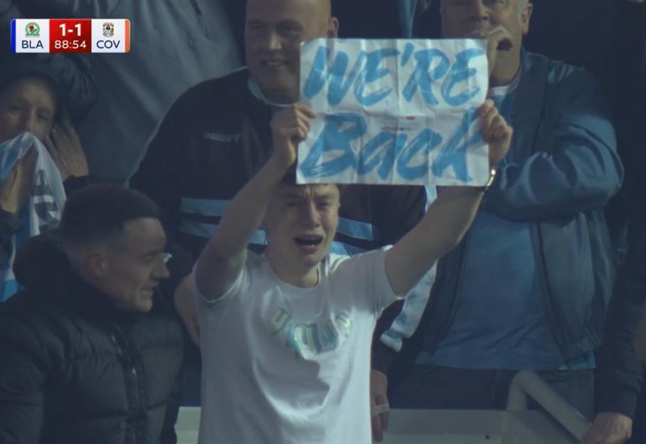 A young fan holds up a "WE'RE Back" sign during a football match.