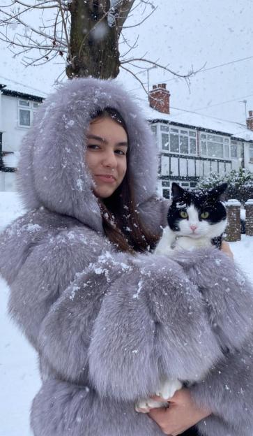 A young woman in a fur-trimmed coat holds a black and white cat while standing in the snow.
