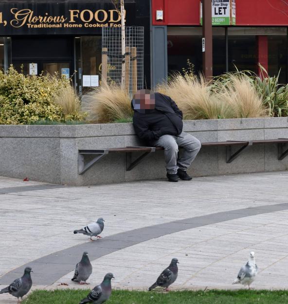 A person in a black jacket and gray jeans sits on a bench in front of a "Glorious FOOD" restaurant, with pigeons on the ground in the foreground.