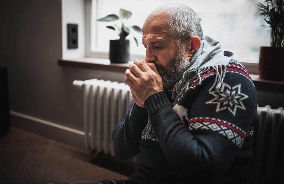A man in a sweater and scarf sits by a radiator, blowing on his hands to keep warm.