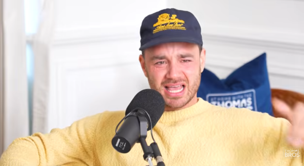 Man wearing a navy baseball cap and yellow sweater, speaking into a microphone with an emotional expression.