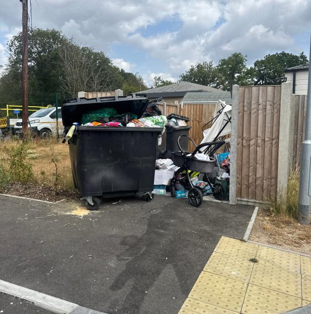 A black overflowing rubbish bin next to a baby stroller filled with bags, alongside a wooden fence at the Four Houses Corner traveller site.