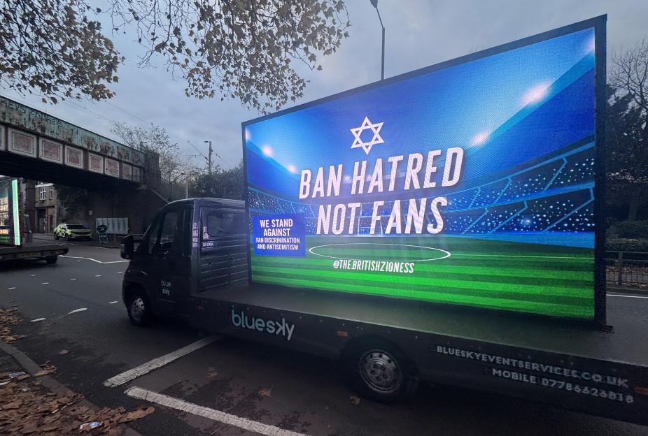 An advertising van displays a message in front of a stadium background reading "Ban Hatred Not Fans" with a Star of David and "We Stand Against Fan Discrimination And Antisemitism."