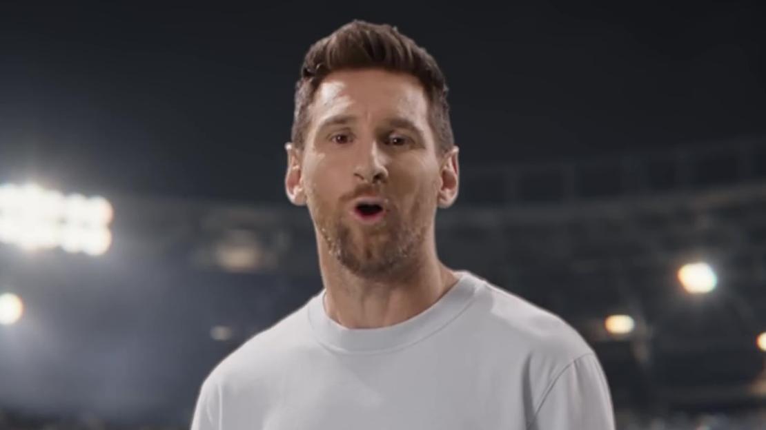 Lionel Messi in a light grey T-shirt, singing in a brightly lit stadium.