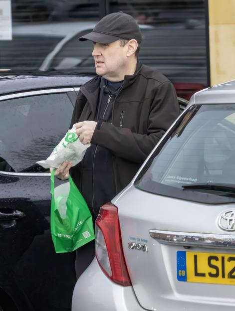 Peter Kay holding a white and a green plastic bag next to a parked car.