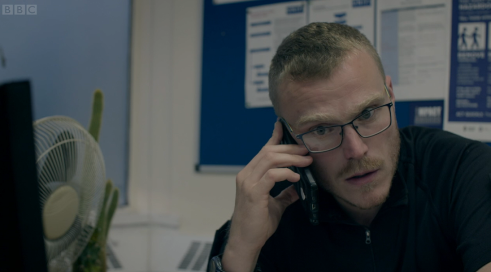 A man with glasses and a beard speaks on a cell phone, looking at a computer screen.