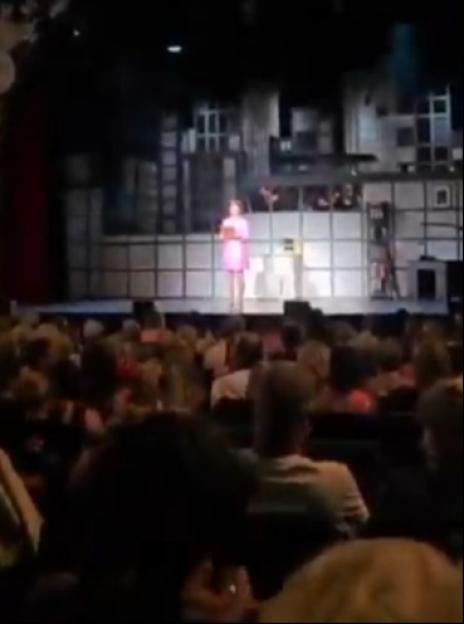 A woman in a pink dress on stage addresses an audience at the 9 to 5 The Musical.