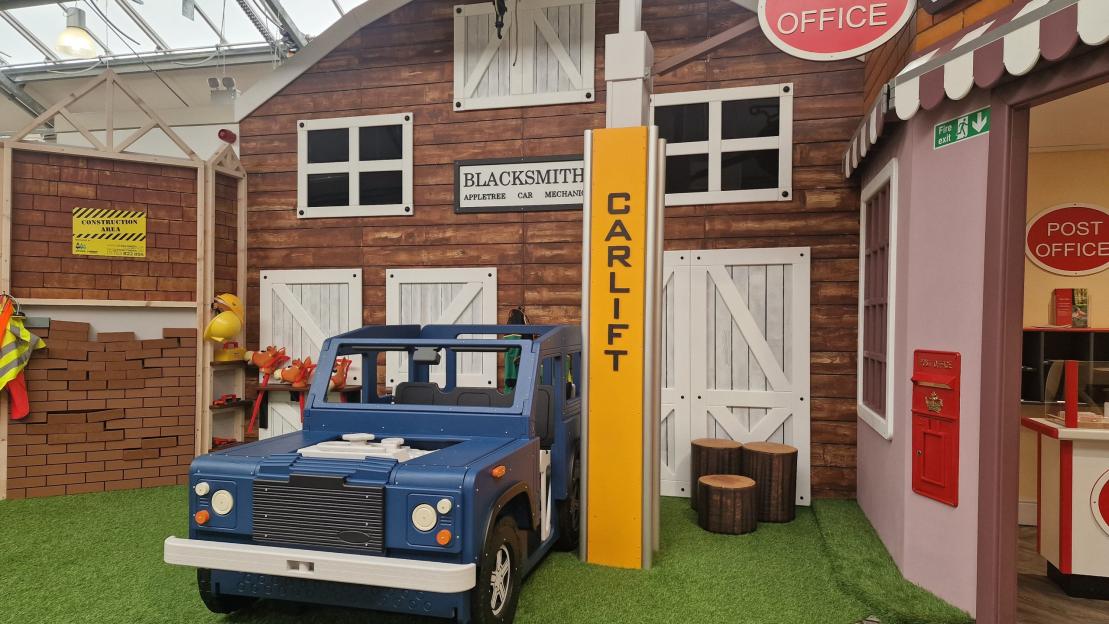 Play area with a blue toy car, a blacksmith building, a construction area, and a post office.