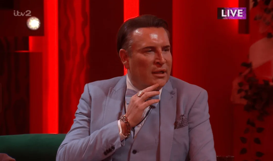 A man in a light blue suit and bow tie speaks on a live television broadcast.
