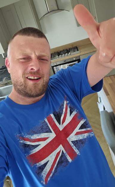 Astron Inman in a blue t-shirt with a faded Union Jack flag gives a thumbs-up.
