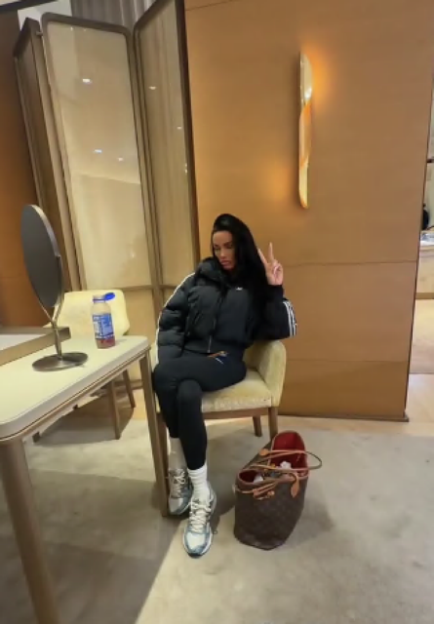 Katie Price's daughter Princess sits on a chair wearing a black puffer jacket, leggings, and sneakers, making a peace sign.