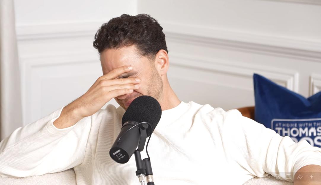 A man in a white sweater covering his face with his hand while speaking into a microphone during a podcast titled "Adam's TRUTH Of What Really Happened In The Jungle... | EP. 45".