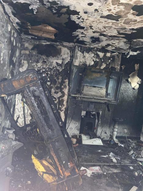A fire-damaged room with blackened walls and ceiling, a charred bed frame, and debris on the floor.