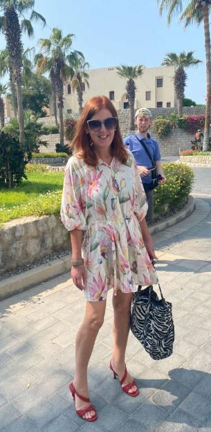 A smiling Shona Sibary wearing a floral dress, sunglasses, and red heels, with a man standing behind her on a sunny street lined with palm trees.