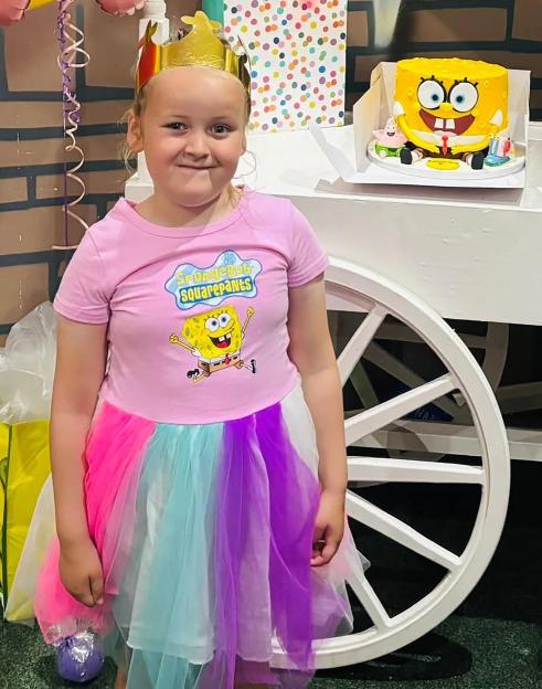 A young girl wearing a gold crown and a pink Spongebob Squarepants shirt and a rainbow-colored skirt stands next to a Spongebob cake on a white cart.