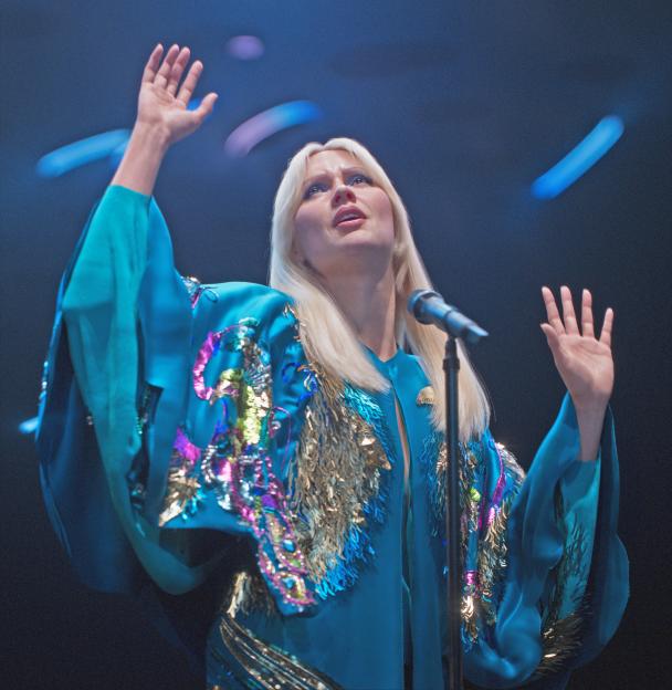 ABBA Voyage concert avatar of a blonde woman in a teal outfit with sequined and feathered accents.