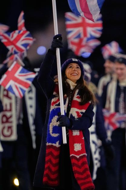 Lilah Fear, Britain's flag bearer, smiles while carrying a flag at the 2026 Winter Olympics opening ceremony.