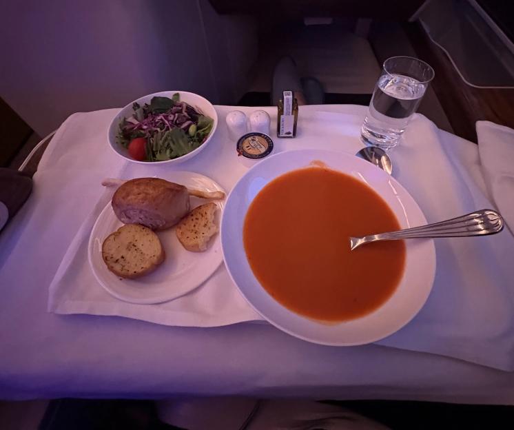 Meal served in a tray in a cabin setting, featuring a bowl of red soup with a spoon, a side salad, bread, a glass of water, and condiments.