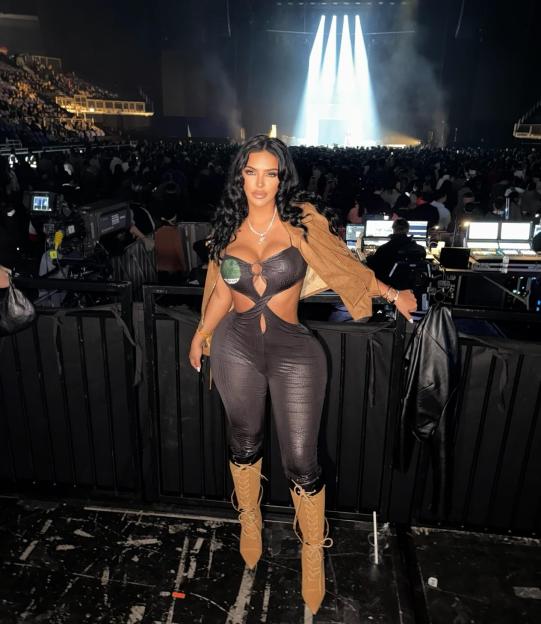 A woman in a black cutout jumpsuit, brown jacket, and tan boots poses in front of a concert crowd and brightly lit stage.