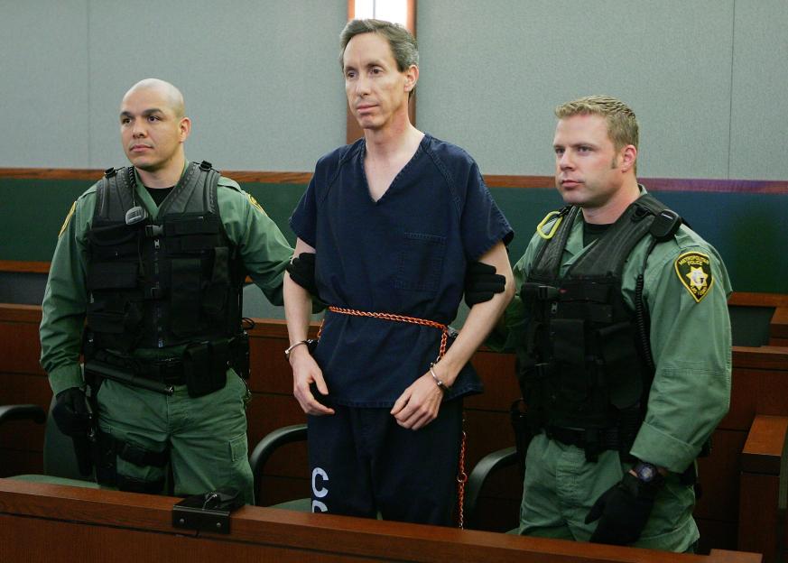 Warren Jeffs (center), wearing an orange belt and handcuffs, between two police officers in green uniforms.