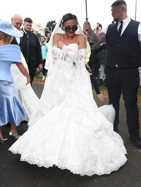 Venezuela Fury arriving at her wedding ceremony in a white lace dress with a long train, accompanied by people holding umbrellas and assisting with her dress.